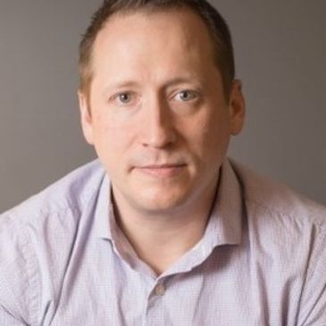 A man with short hair wearing a light purple shirt looks at the camera.