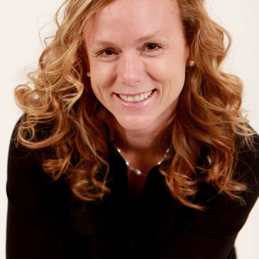 Smiling woman with curly red hair wearing a black top and pearl necklace.