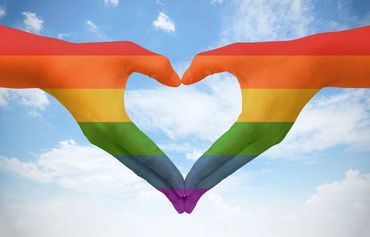 Hands forming a heart with a rainbow flag overlay against a blue sky.
