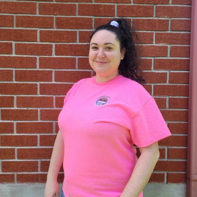 Femme souriante portant un t-shirt rose devant un mur en briques rouges.