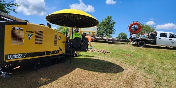 Workers operating a yellow drilling machine on a grassy field under a blue sky.