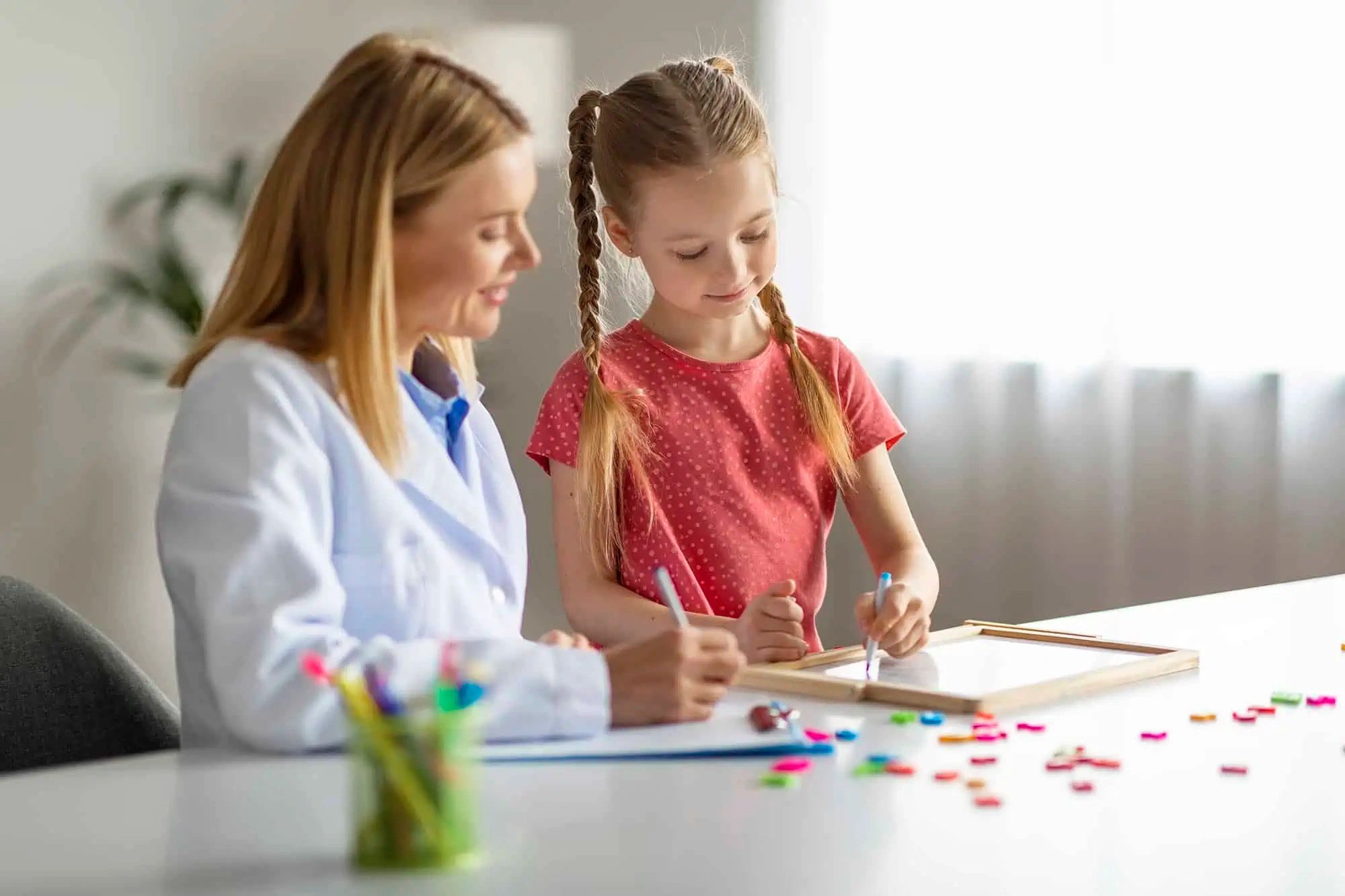 Female teacher helping young girl with homework at a desk.