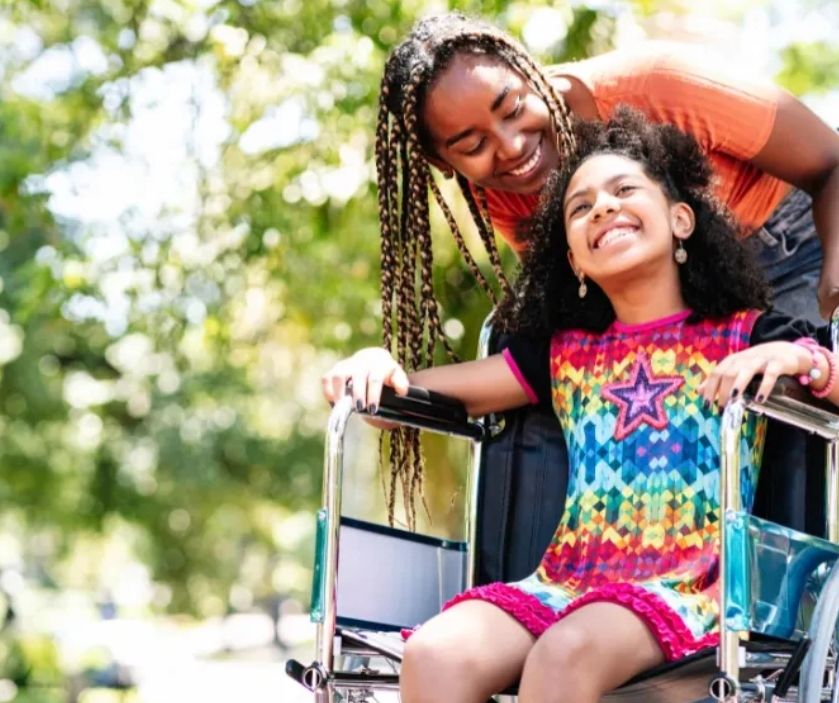 A smiling young black women in long braids pushing the wheelchair of a happy little girl 