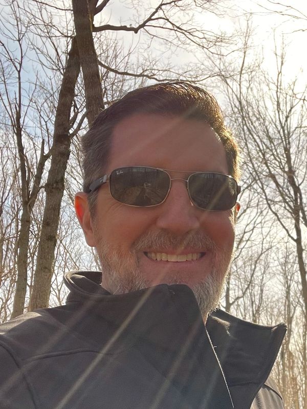 Smiling man wearing sunglasses in a sunlit forest with bare trees.