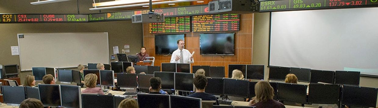 Classroom setting with stock market data screens and a speaker.