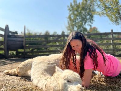 A woman in a pink shirt lies beside a resting alpaca on straw.