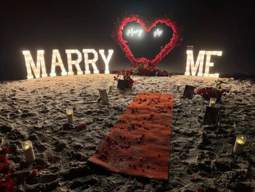 Romantic beach proposal setup with illuminated 'Marry Me' signs and rose decorations.