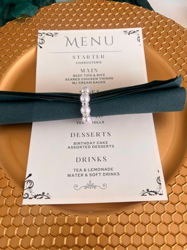 Elegant menu on a gold plate with a teal napkin and pearl napkin ring.