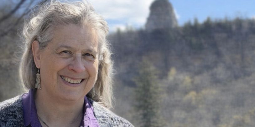 Smiling woman with gray hair outdoors with a blurred forest background.