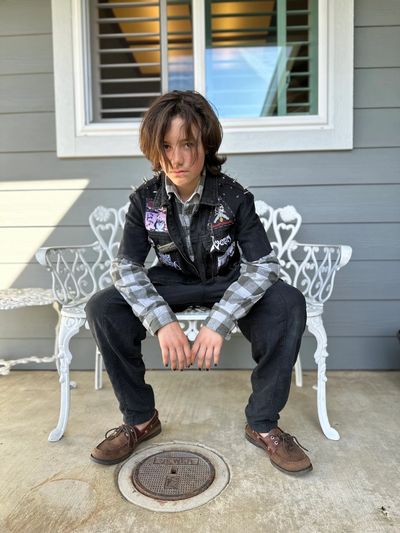 Teen boy in layered punk rock outfit sitting on white bench outdoors.