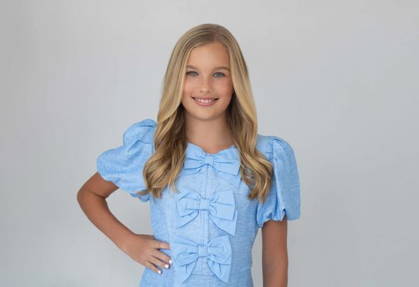 A smiling girl in a blue dress with bows.