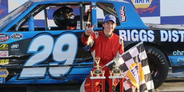 Race car driver celebrating victory with trophies and checkered flag.