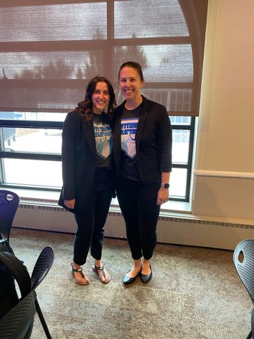 Two women smiling indoors wearing matching shirts and black blazers.