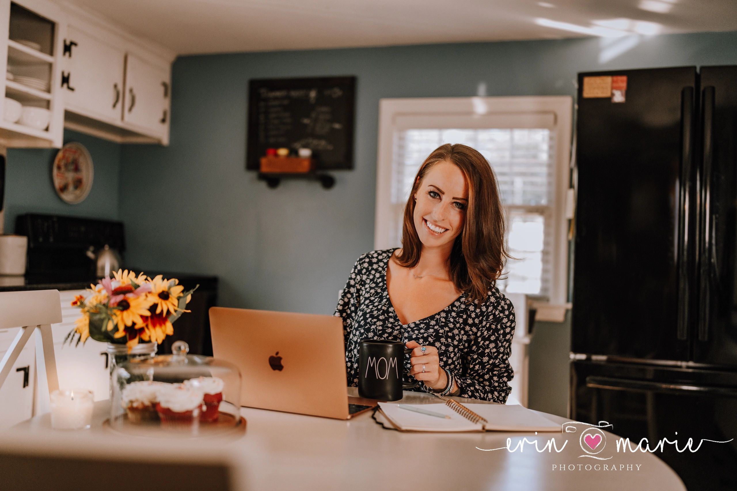 Smiling woman holding a 'MOM' mug in a cozy kitchen with laptop and flowers.