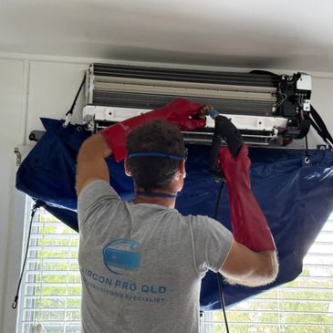 Technician cleaning a wall-mounted air conditioner with protective gloves and equipment.