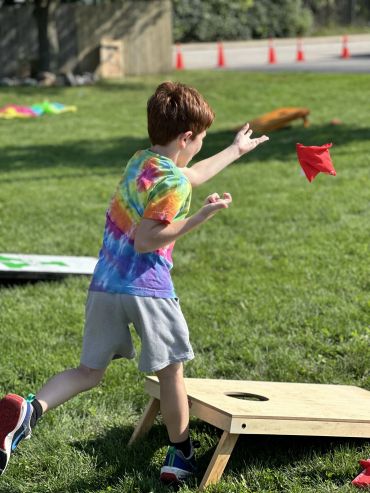A boy in a tie-dye shirt tossing a red bean bag towards a cornhole board.