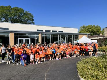 Large group of walk participants in orange shirts posing outdoors in front of a building on a sunny day.