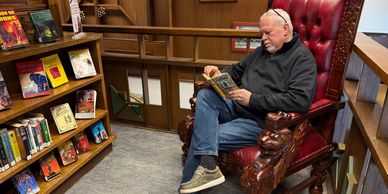 Man reading a book in a large, ornate wooden chair in a library corner.