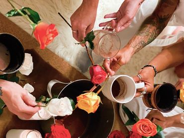 People holding roses and mugs around a pot on a table.