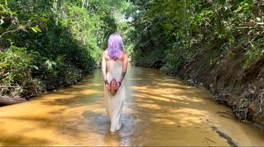 Person with purple hair standing in a shallow river surrounded by dense greenery.