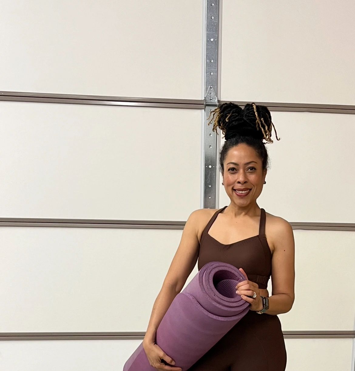 Woman in brown workout clothes holding a purple yoga mat, smiling.