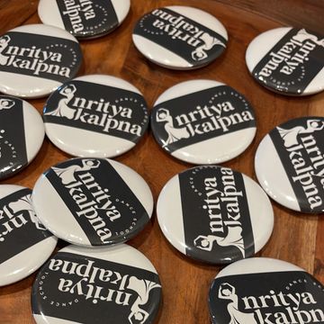 Round badges with 'Nritya Kalpna' logo scattered on a wooden surface.