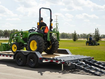 A green tractor is loaded onto a trailer with ramps on a sunny day.