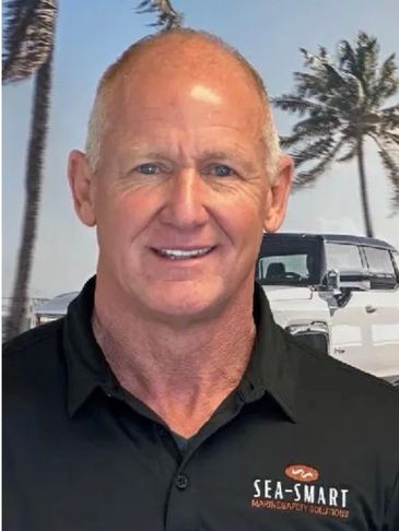 Smiling man in a black Sea-Smart shirt with palm trees and a white vehicle in the background.