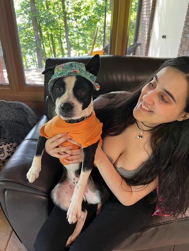 Smiling woman holding a dressed-up dog on a couch near a window.