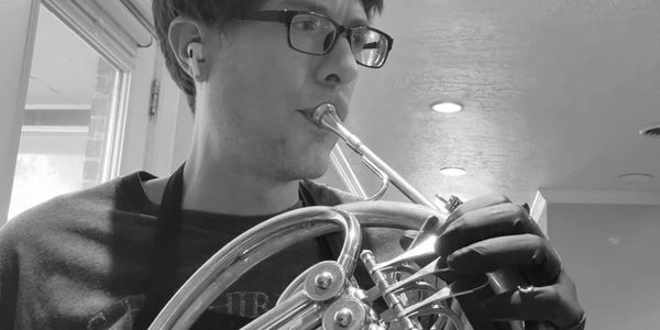 Man playing a French horn indoors, wearing glasses and earbuds.