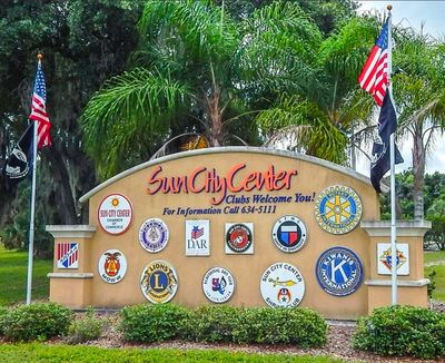 Welcome sign for Sun City Center with various club logos and two American flags.