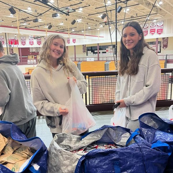 Two women smiling while holding bags in a gymnasium.