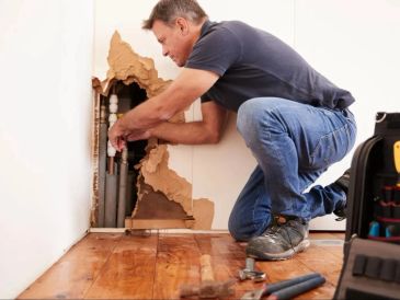 Man repairing pipes inside a damaged wall.