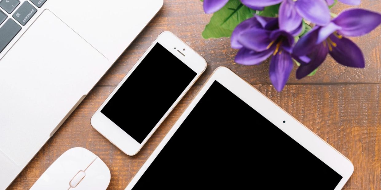 laptop, mouse, cell phone and tablet arranged on wooden surface with a purple iris bouquet.