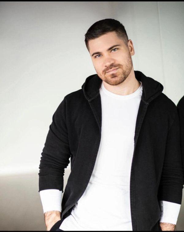 Young man in a black hoodie and white shirt posing casually indoors.