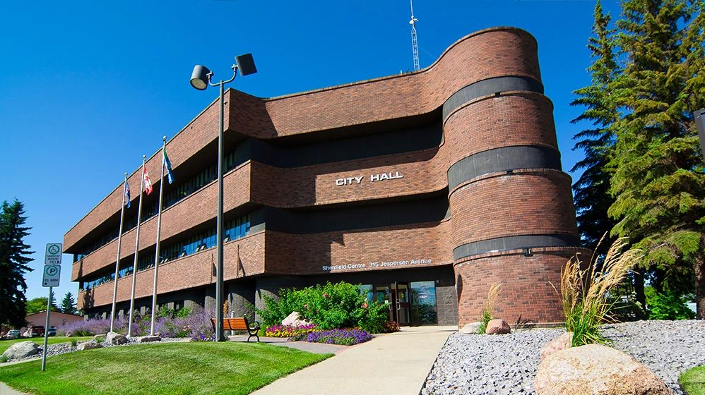 Modern brick city hall building with flags and landscaped garden under clear blue sky.