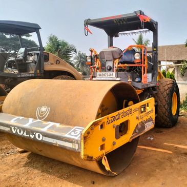 A decorated Volvo road roller parked on a dirt ground with palm trees in the background.