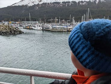 A child in a blue knit hat gazes at a marina filled with boats and surrounded by forested mountains.