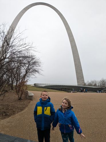Two children stand near the Gateway Arch on a cloudy day.