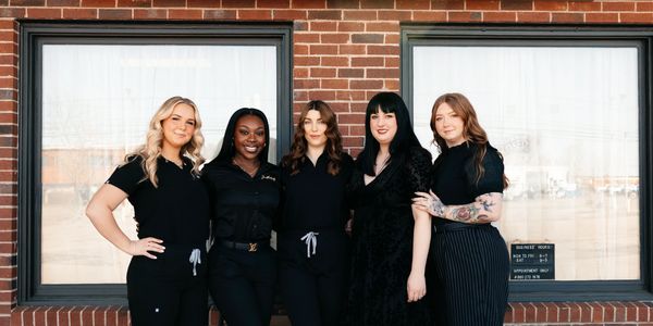 Five diverse women posing confidently outside a brick building with large windows.