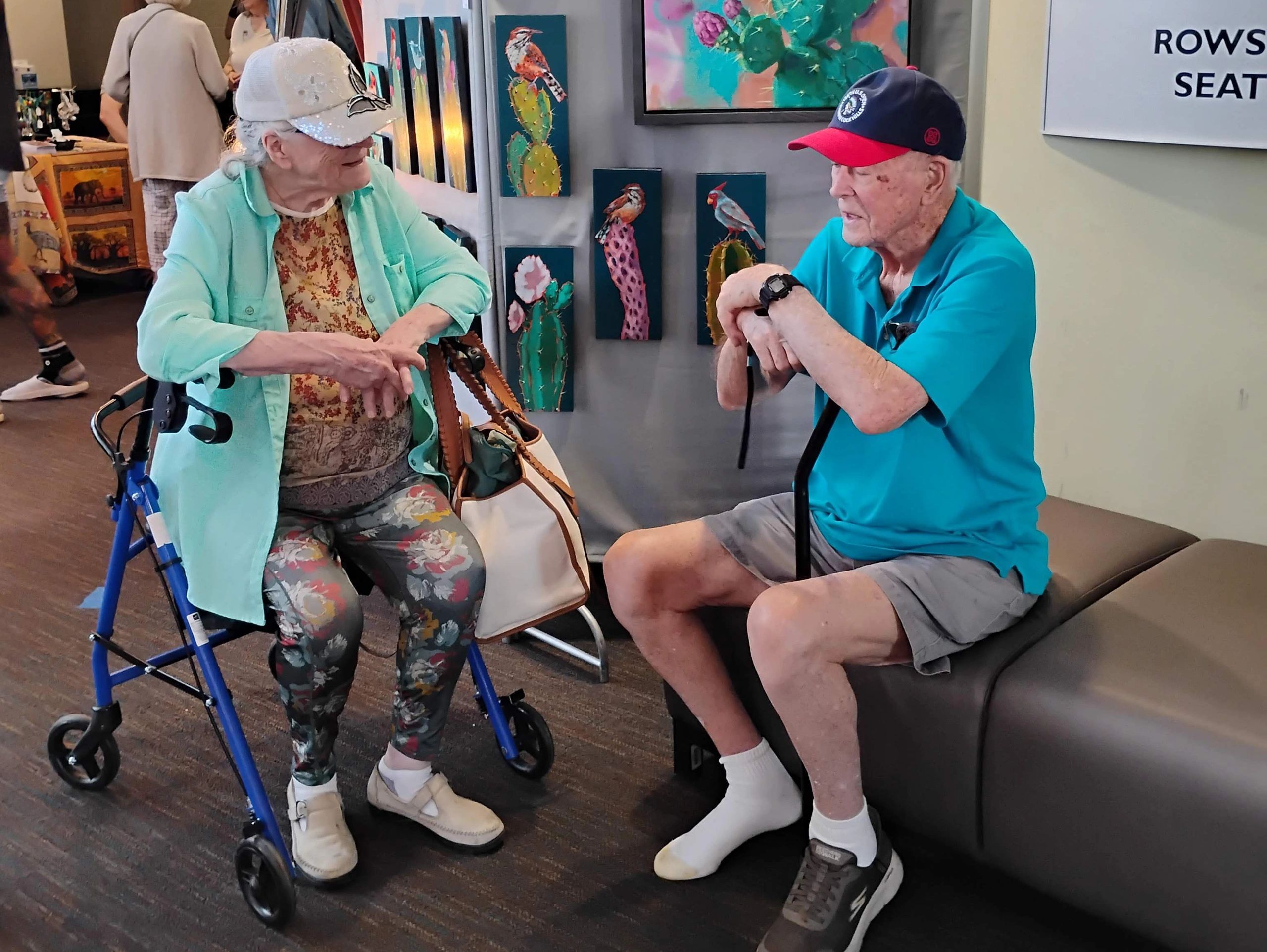 Two elderly people seated in front of hummingbird inspired paintings.