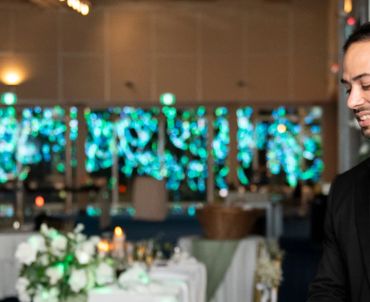 A smiling waiter in black attire at an elegantly decorated dining area with floral arrangements and blue lights.