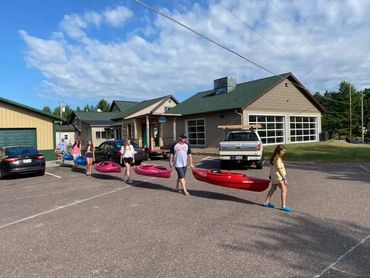 Group carrying kayaks in a parking lot near buildings under a blue sky.