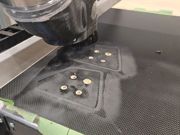 CNC machine carving holes into a carbon fiber sheet with dust and tape around.