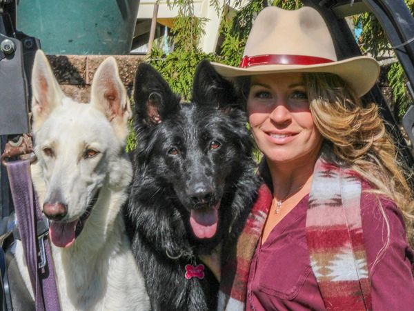 Woman in hat posing with two German Shepherd dogs, one white and one black.