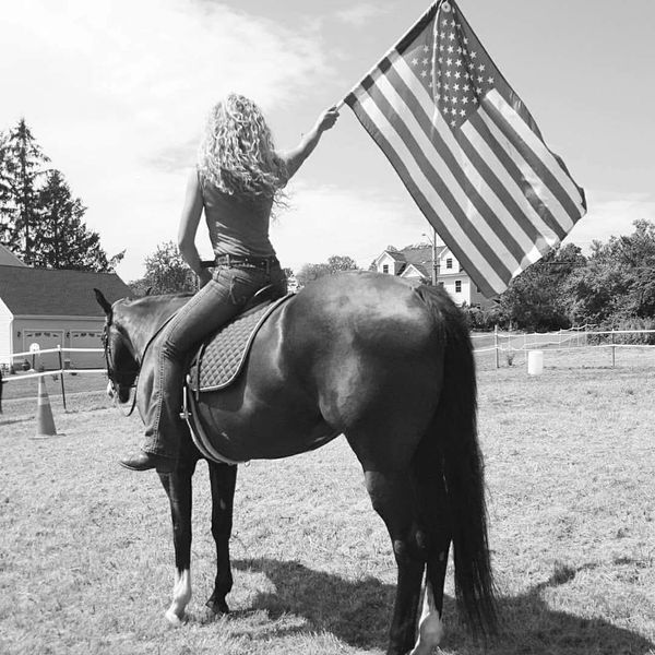 A woman on horseback facing away from the camera, she hold a large American flag in the air