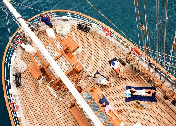 Four people relaxing on the deck of a wooden sailboat in clear blue water.