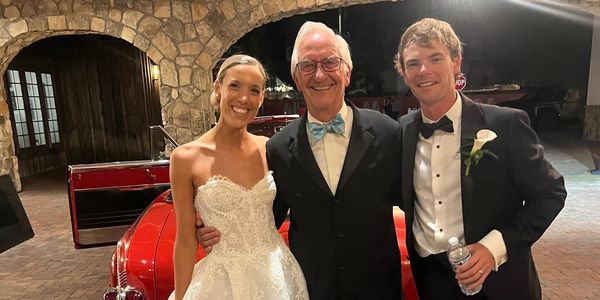 Bride and groom pose with an older man in front of a red vintage car.