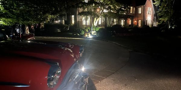 Vintage red car with headlights on in front of a warmly lit mansion at night.