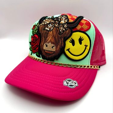 Colorful trucker hat with embroidered cow, smiley face, rose, and chain detail.